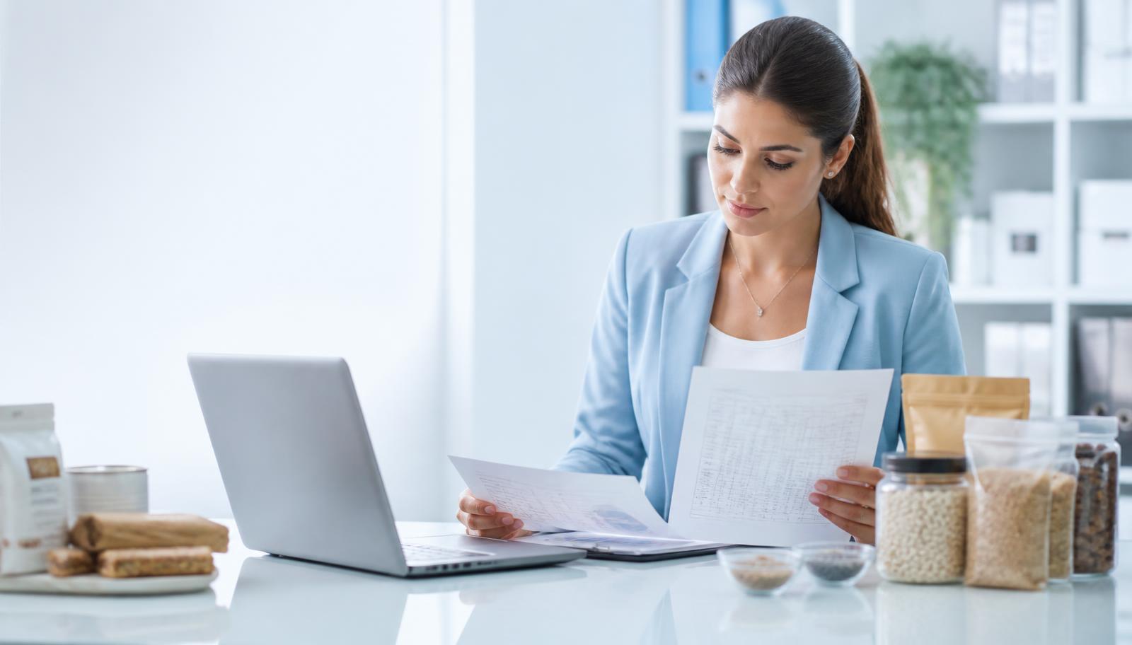 Regulatory affairs specialist reviewing technical product documentation and specification sheets beside packaged food products and ingredient samples